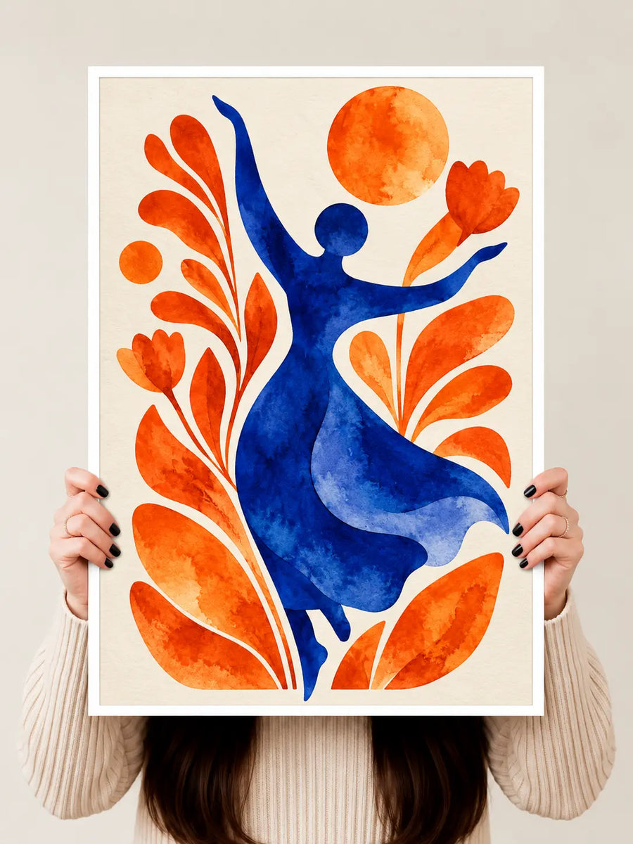 A framed art print featuring a stylized blue dancer surrounded by orange leaves and a sun on a cream background, held up by a person wearing a beige ribbed sweater against a light gray wall.