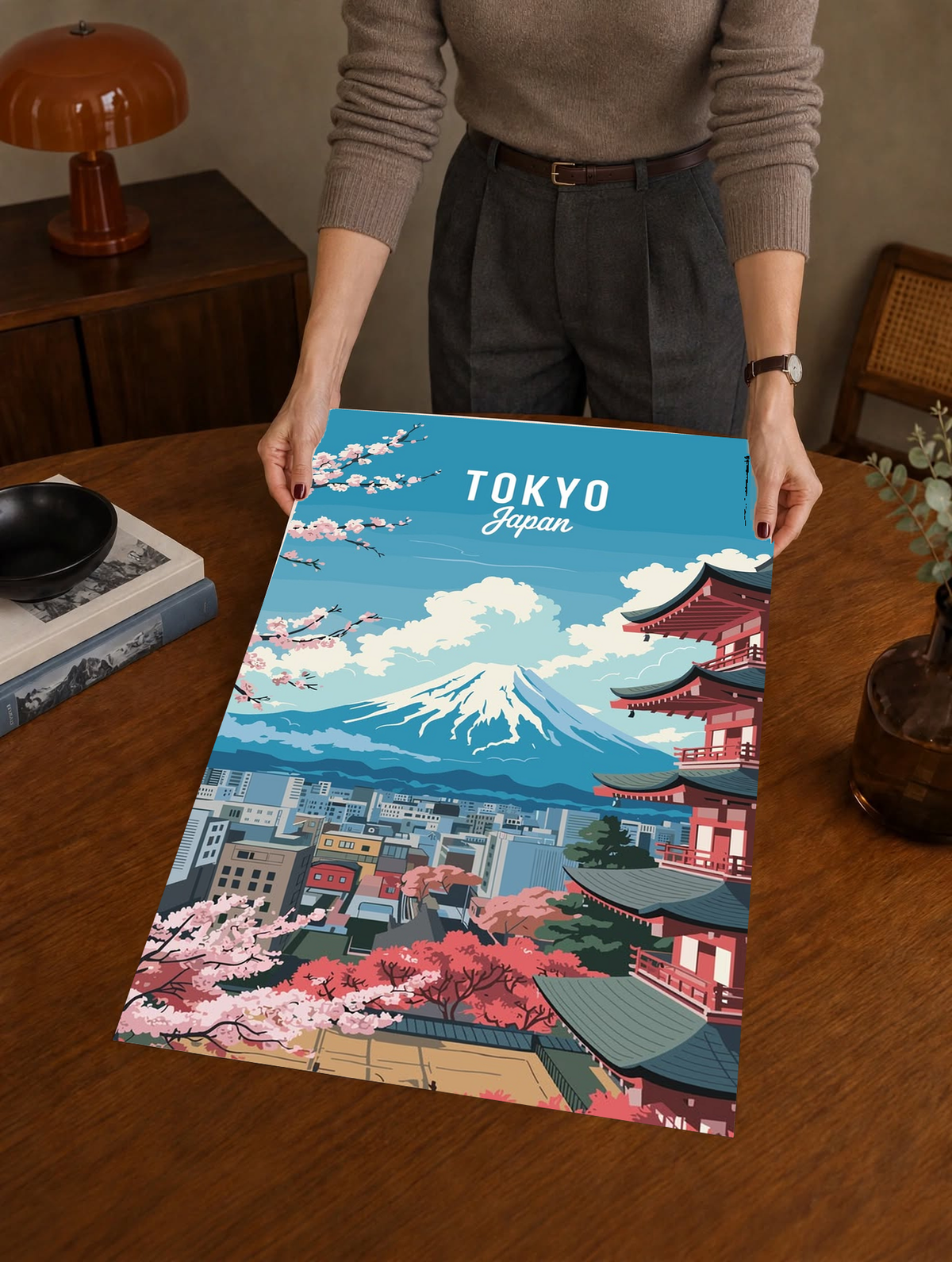Japanese Cherry Blossoms Wall Art held upright by a person above a wooden table, presenting a colourful Tokyo scene with Mount Fuji, a tiered red pagoda and flowering pink branches.