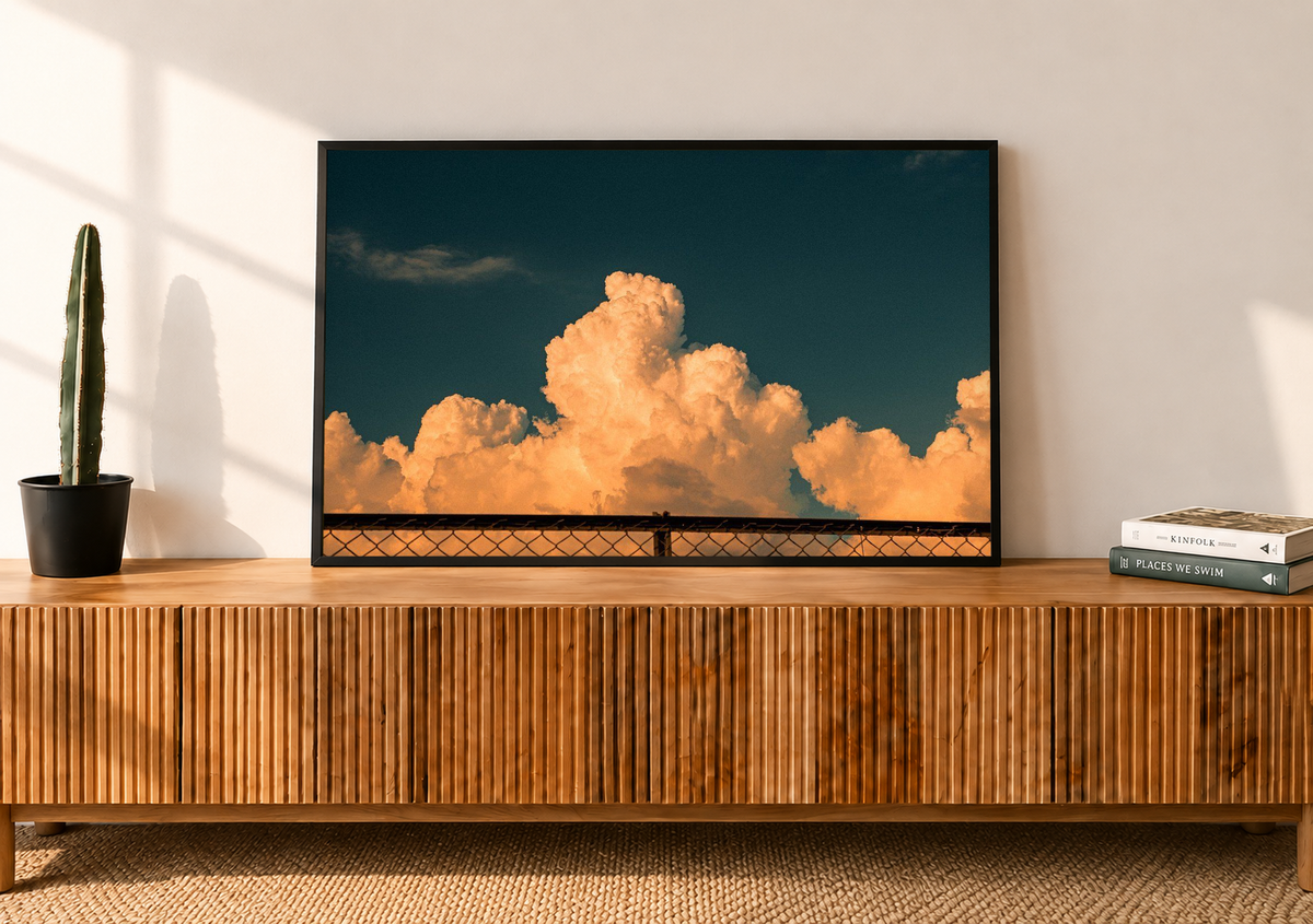 Orange Cloud Sky Wall Art displayed in a slim black frame above a ribbed wooden console, with glowing peach clouds beneath a deep teal sky and afternoon sunlight crossing the neutral living room.