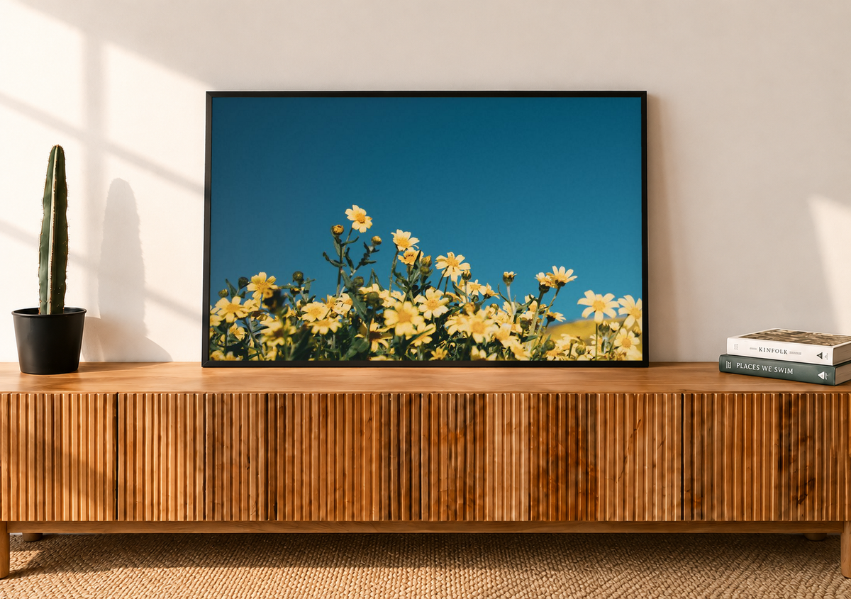 Black framed yellow wildflowers print displayed above a wooden console, with golden flowers and a deep blue sky adding color to a sunlit living room.
 Copy Alt Text