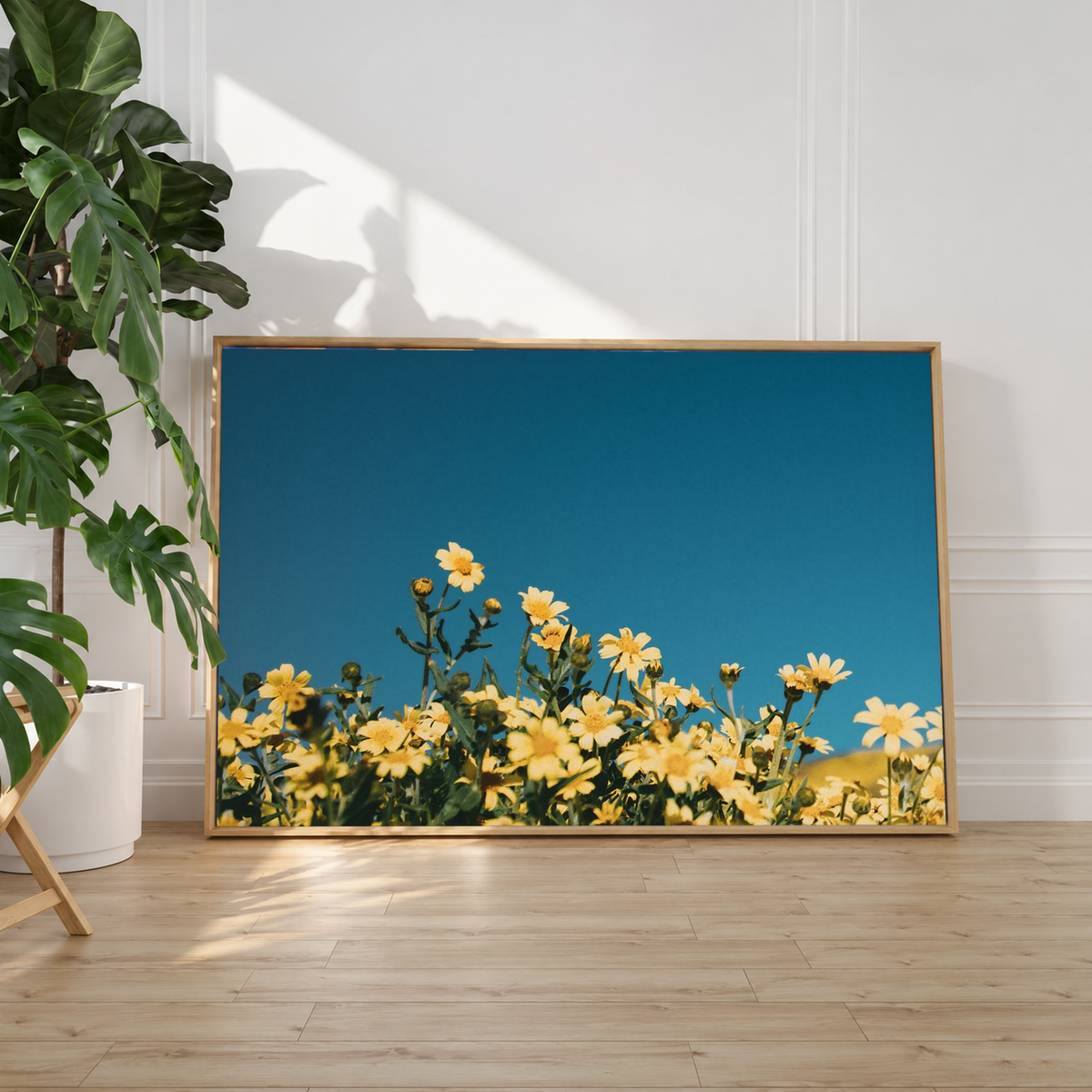 Yellow floral wall art leaning beside a leafy indoor plant, with rich blue tones contrasting against the room's soft neutral decor.
 Copy Alt Text