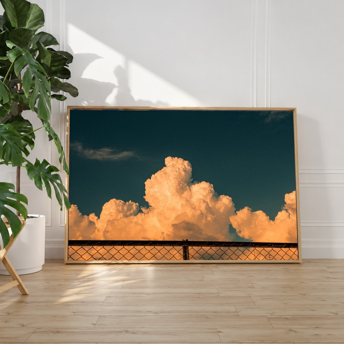 Orange cloud artwork resting against layered white pillows and soft bedding, its dark blue sky and sunlit cloud formations creating a warm visual contrast within the relaxed bedroom setting.