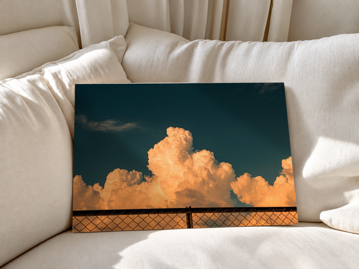Unframed landscape cloudscape print showing sunlit apricot clouds rising above a metal fence, contrasted with a wide expanse of dark blue-green sky and subtle distant cloud formations.