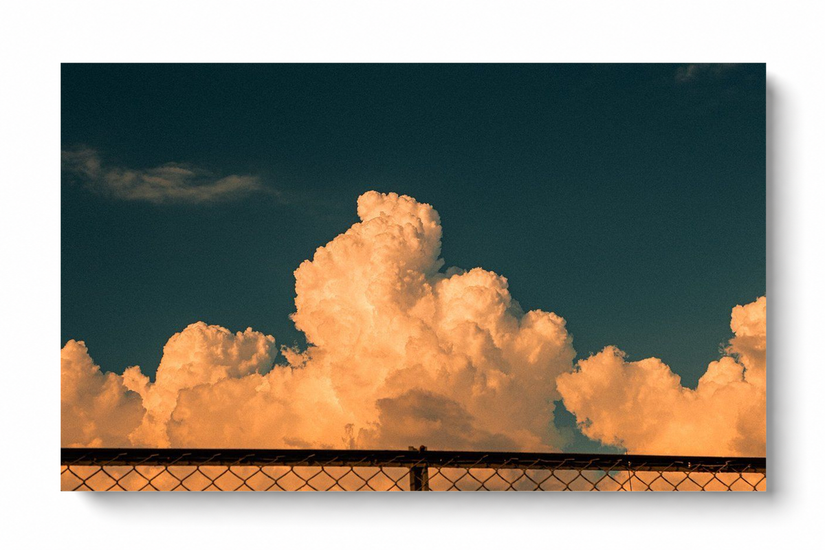Horizontal orange cloud sky poster featuring towering peach-colored clouds against a deep teal sky, with a dark chain-link fence stretching across the bottom of the atmospheric composition.
