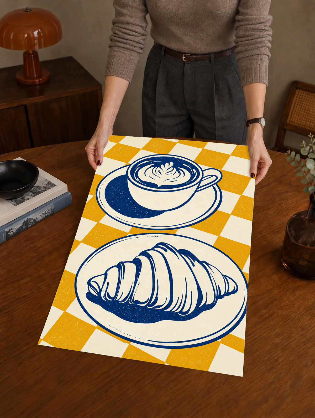 Tazza blu navy con piattino sopra un croissant su un piatto separato, davanti a uno sfondo a quadri senape e crema, come poster senza cornice tenuto sopra un tavolo di legno da una persona in piedi dietro.