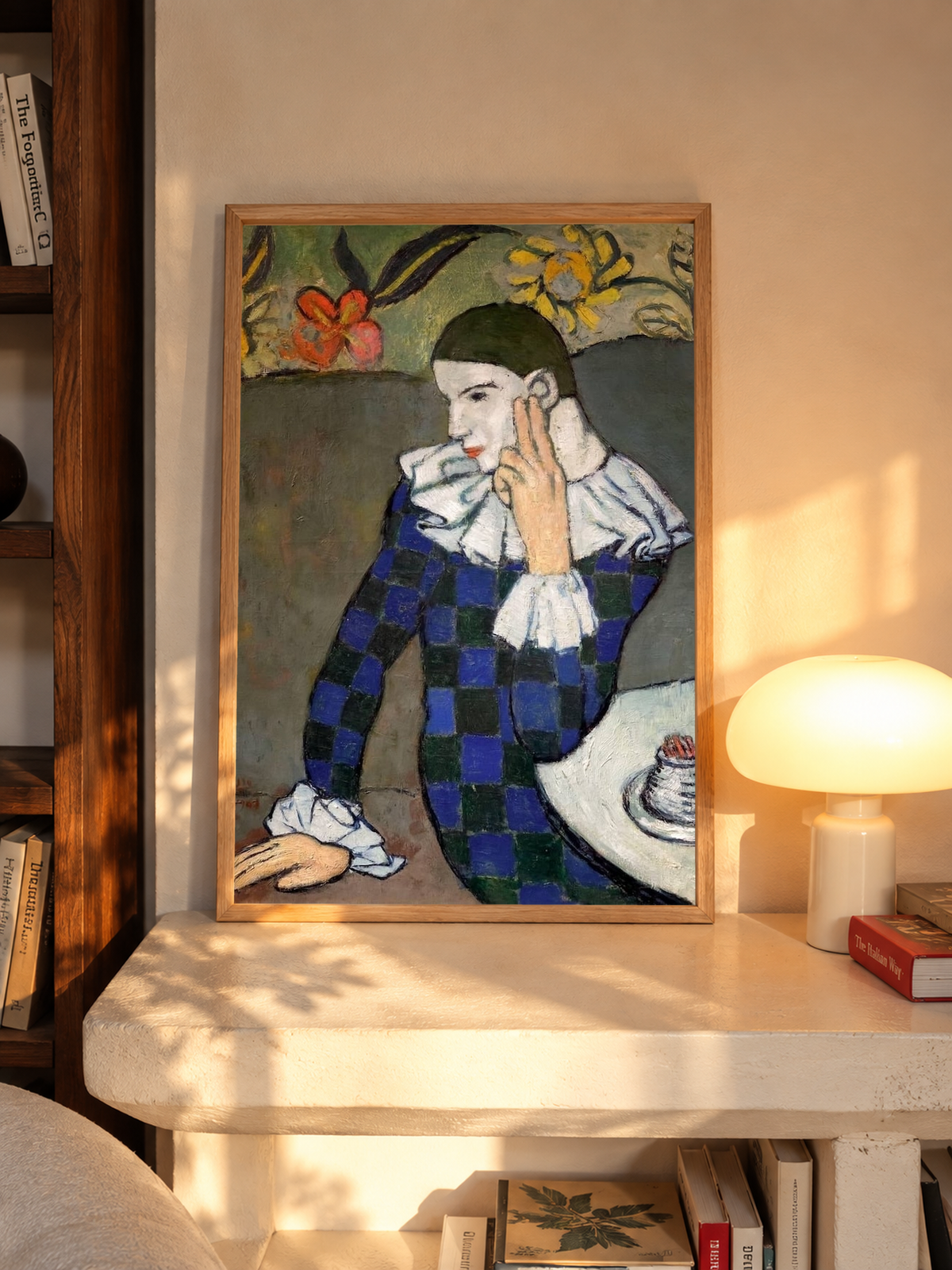 Warm light from a table lamp falls beside the framed Picasso Seated Harlequin Print, highlighting the seated figure, curved cafe table and red-and-yellow flowers above the muted green background.