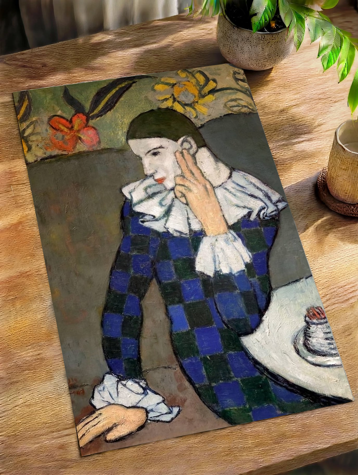 Resting against textured cream chair upholstery, the Picasso Seated Harlequin Print pairs deep blue costume squares with pale ruffles, earthy background tones and a small vessel on the curved cafe table.