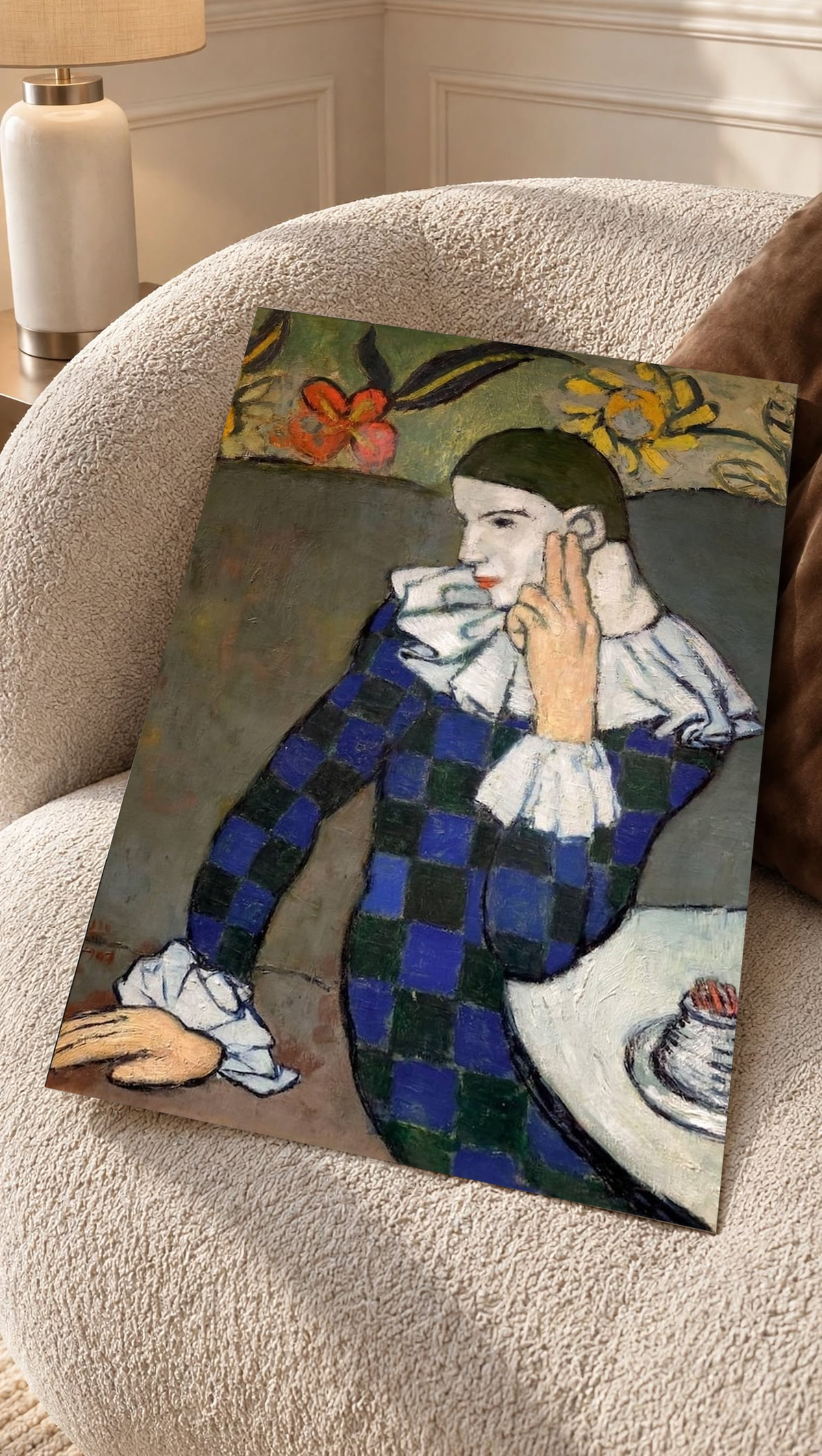 A hand supports the Picasso Seated Harlequin Print against a light wall, offering a close view of the thoughtful facial expression, raised fingers and floral details surrounding the dark-haired figure.