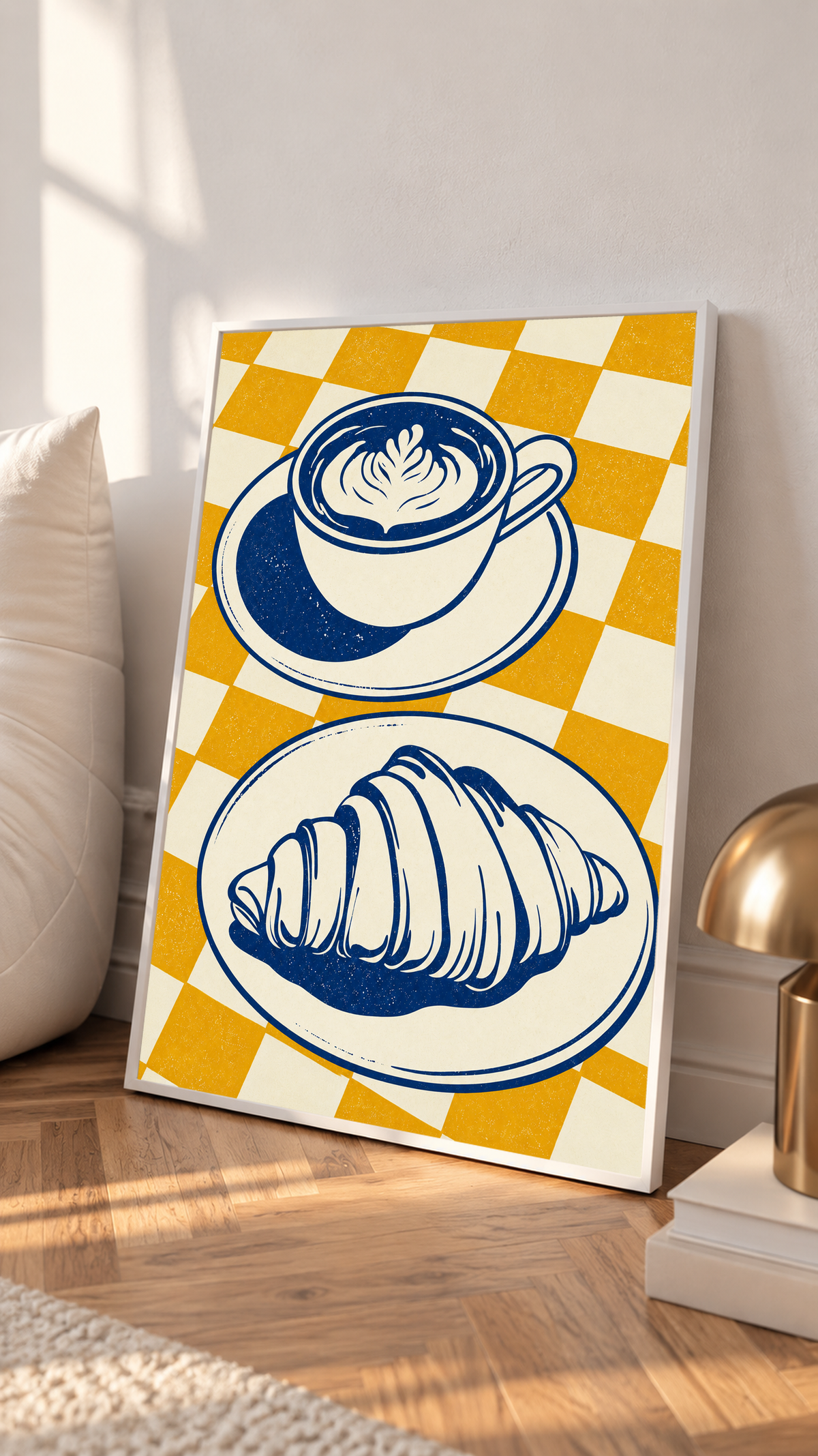 Tazza blu navy con piattino sopra un croissant su un piatto separato, davanti a uno sfondo a quadri senape e crema, in una cornice bianca appoggiata su un pavimento di legno, accanto a un cuscino chiaro e una lampada color ottone.