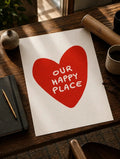 Lámina sobre una mesa de madera: un corazón rojo con el texto blanco OUR HAPPY PLACE sobre fondo crema.