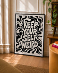 Black-framed black-and-white typographic art print standing on the floor; wavy lines and Keep Yourself Weird text
