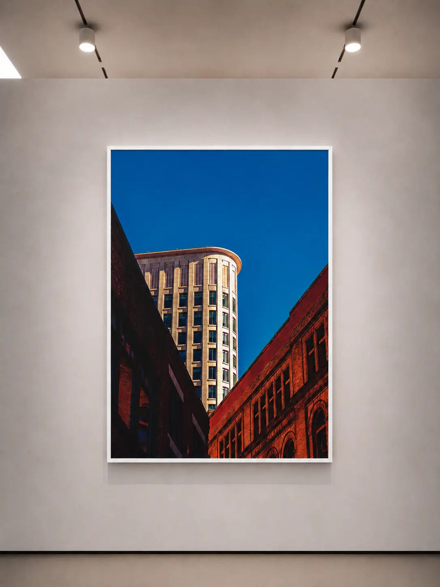 Póster vertical con borde blanco que muestra un edificio alto y una calle de ladrillo rojo bajo un cielo azul