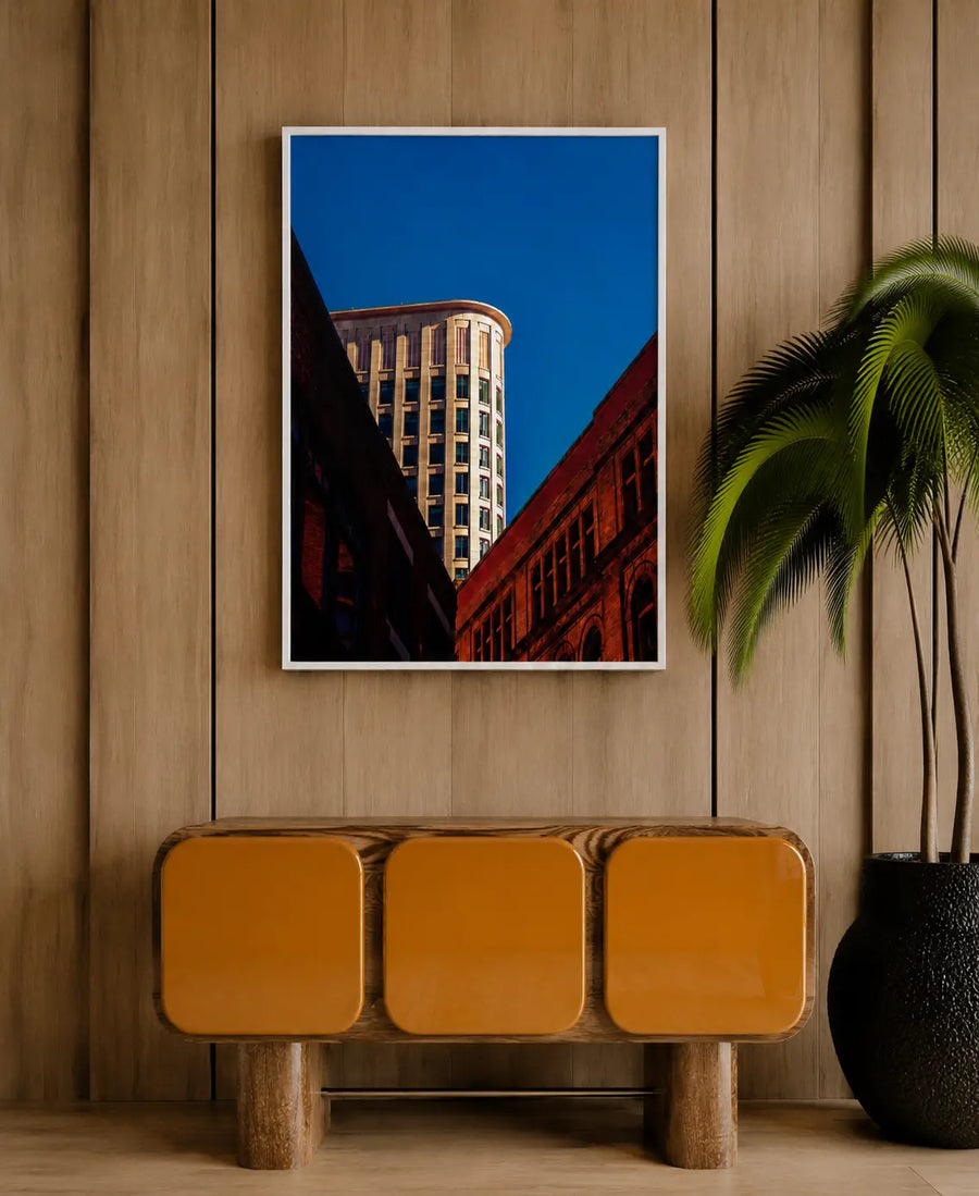 Impresión vertical urbana con marco blanco que muestra un edificio alto, fachadas de ladrillo rojo y cielo azul