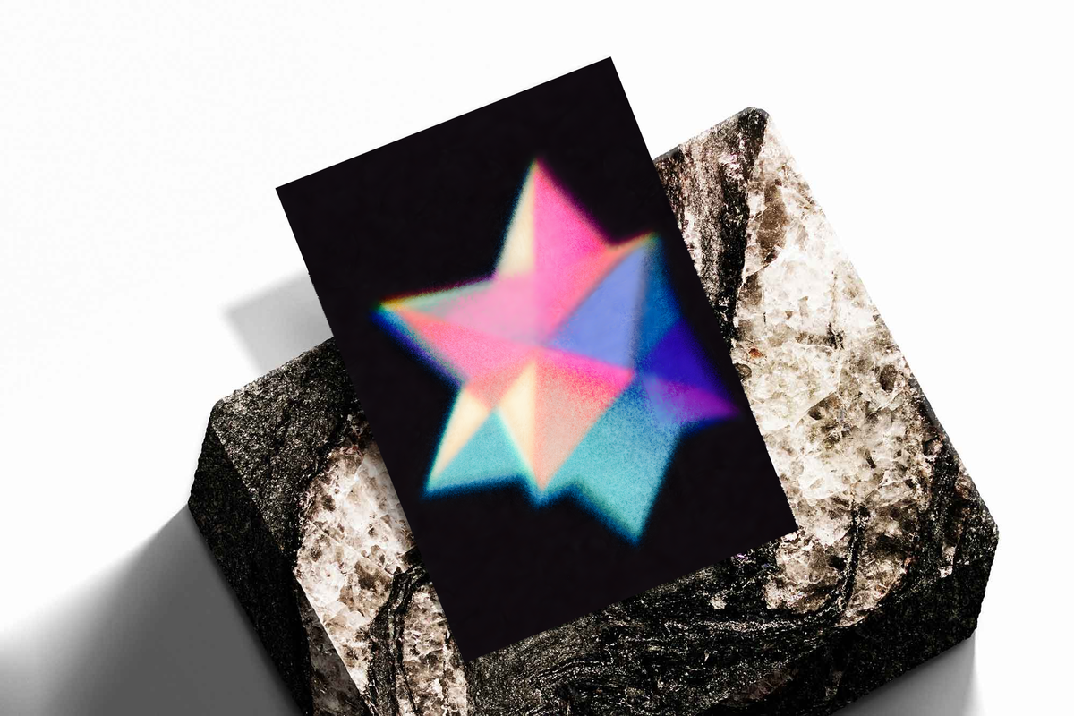 Overlapping pink cyan blue and purple geometric star shapes with grain and soft glow on black, shown as an unframed print tilted on a dark stone display against a light background