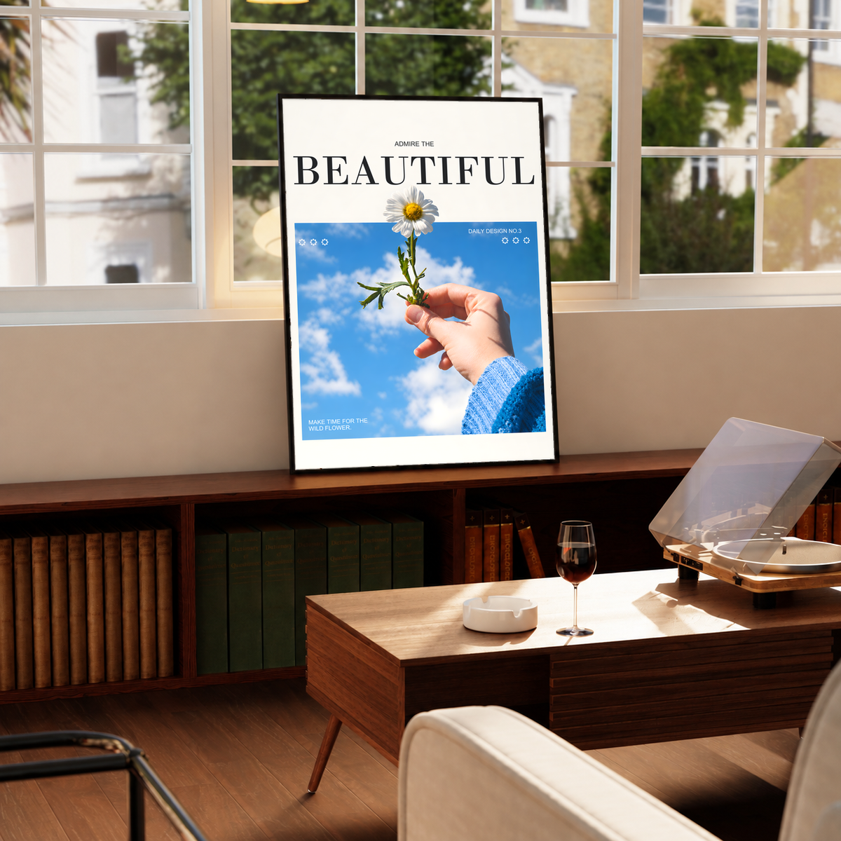 Hand holding a white daisy under blue sky with Admire the Beautiful lettering; framed print beside a window
