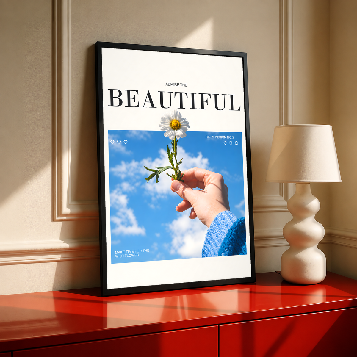 Hand holding a white daisy under blue sky with Admire the Beautiful lettering; black framed print on a red cabinet