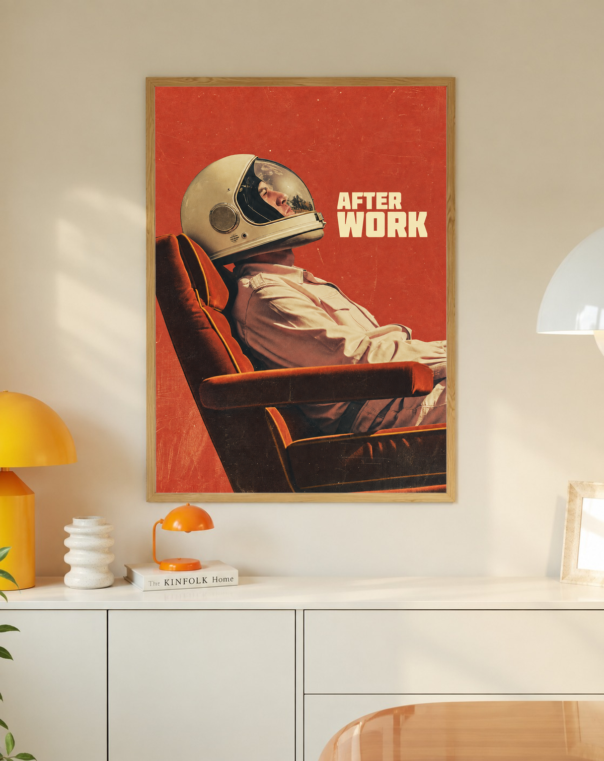 An astronaut reclining in an armchair against a burnt-orange ground in a slim oak frame on a cream wall above a white cabinet