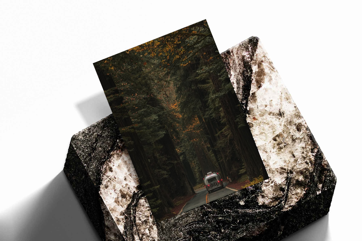 Unframed artwork depicting rear view of a car on a road beneath tall autumn trees, presented diagonally on rough stone.