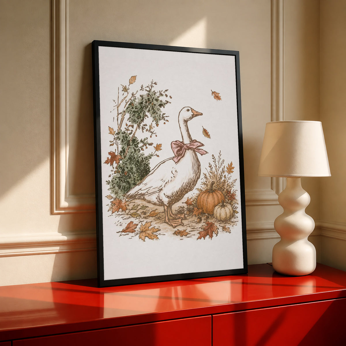 White goose with a rust pink bow among pumpkins and scattered autumn leaves in a black frame on a red cabinet beside a cream lamp