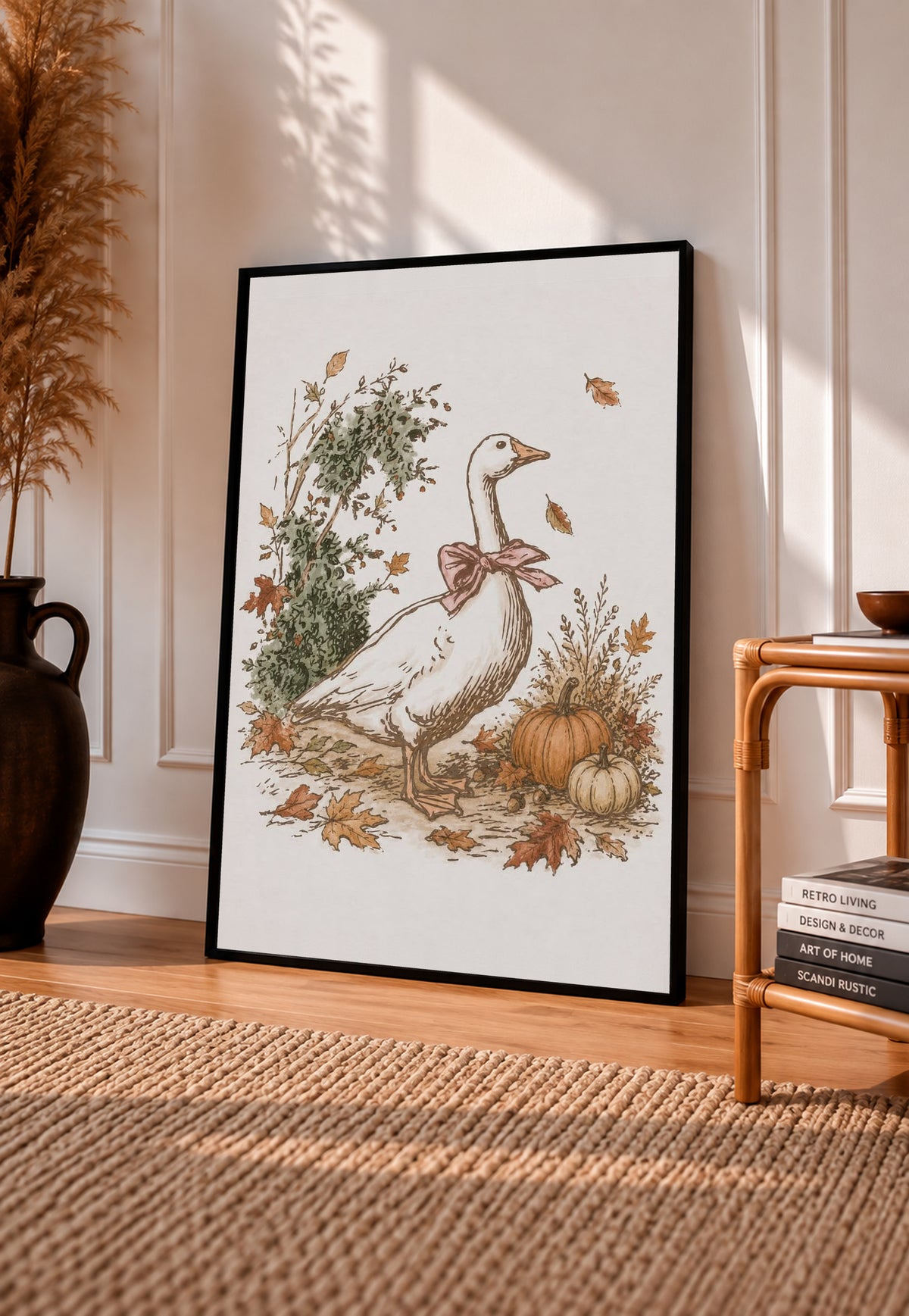 White goose with a rust pink bow among pumpkins and scattered autumn leaves in a black frame leaning on a sunlit wooden floor beside books and a rattan seat