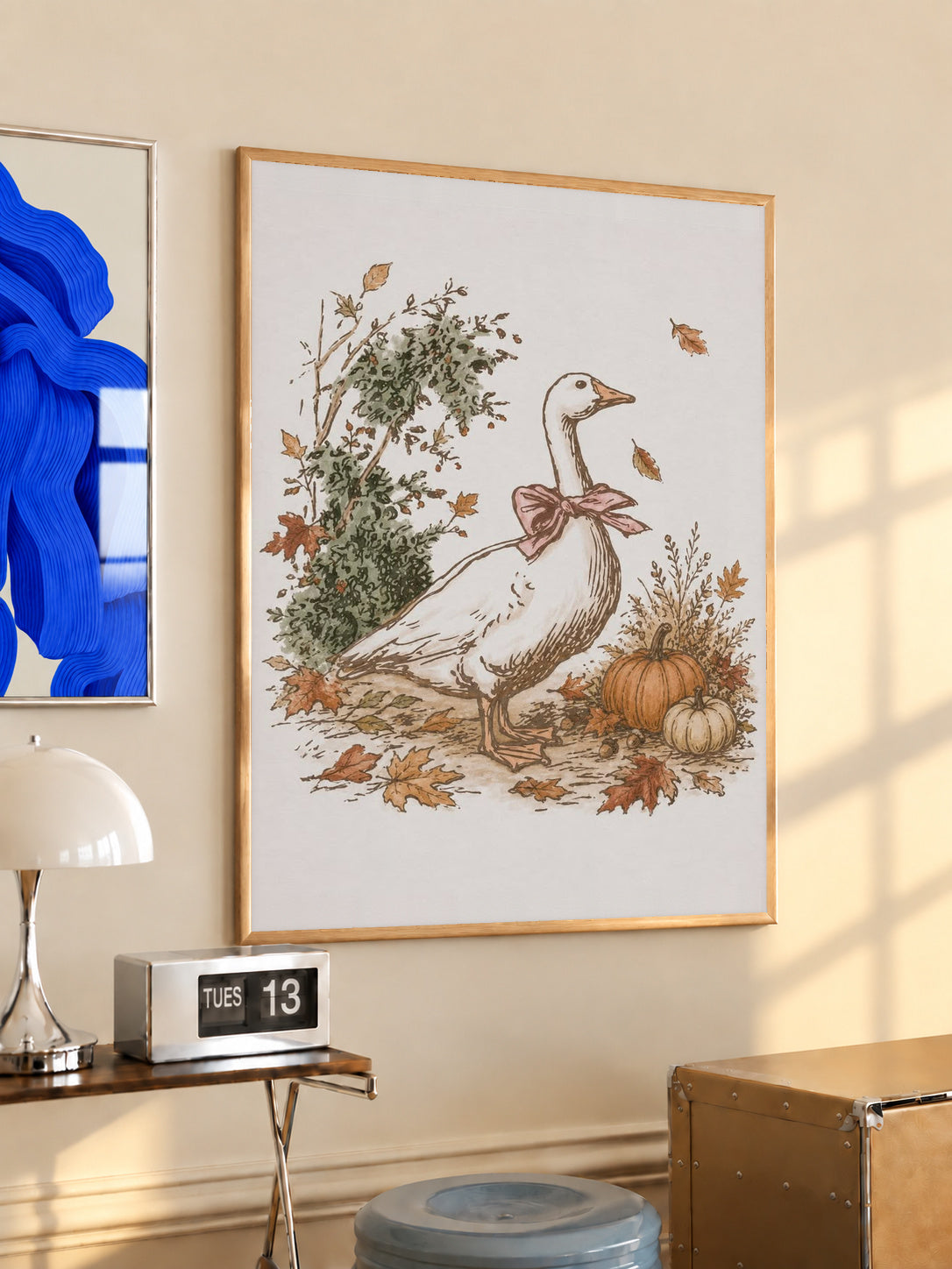 White goose with a rust pink bow among pumpkins and scattered autumn leaves in an oak frame above a console beside a blue print and calendar