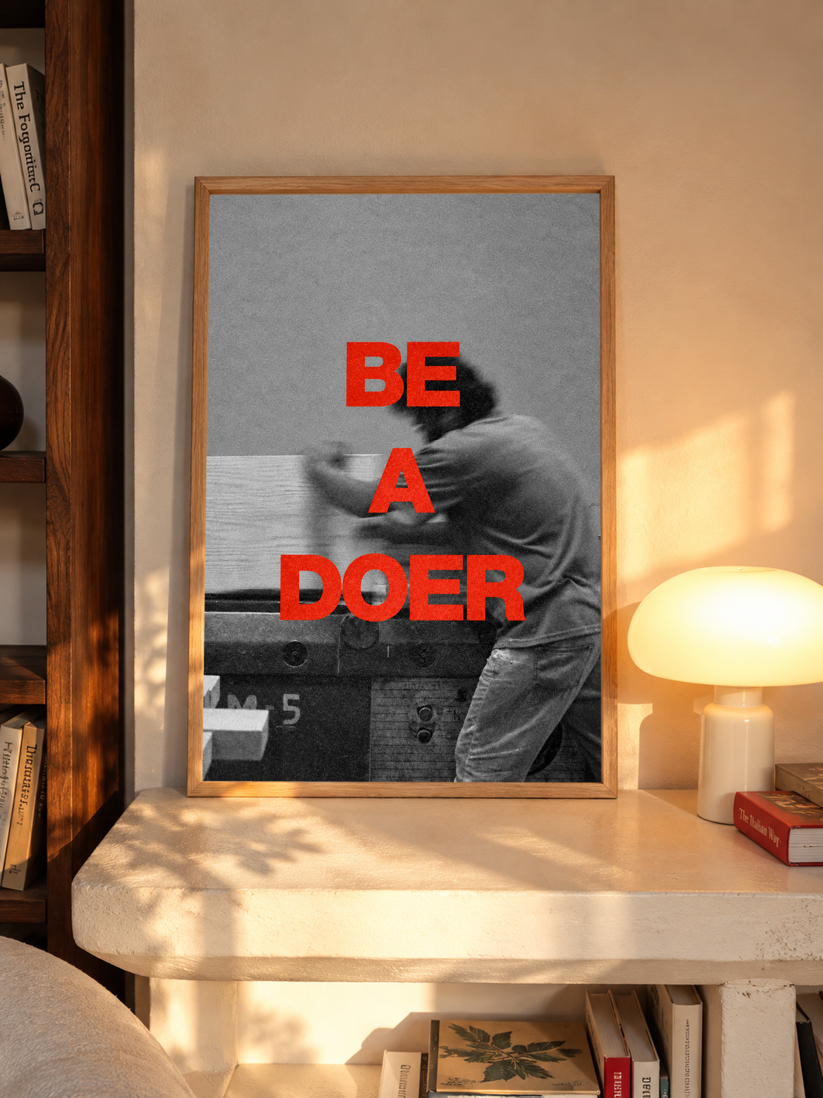 Red Be a Doer lettering over a motion-blurred monochrome figure in an oak frame above a pale wooden sideboard beside a glowing lamp