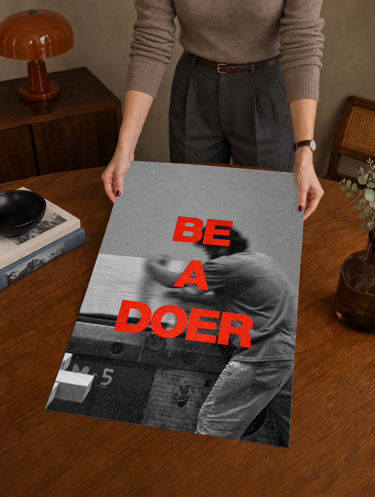 Red Be a Doer lettering over a motion-blurred monochrome figure as a large unframed poster held above a wooden desk