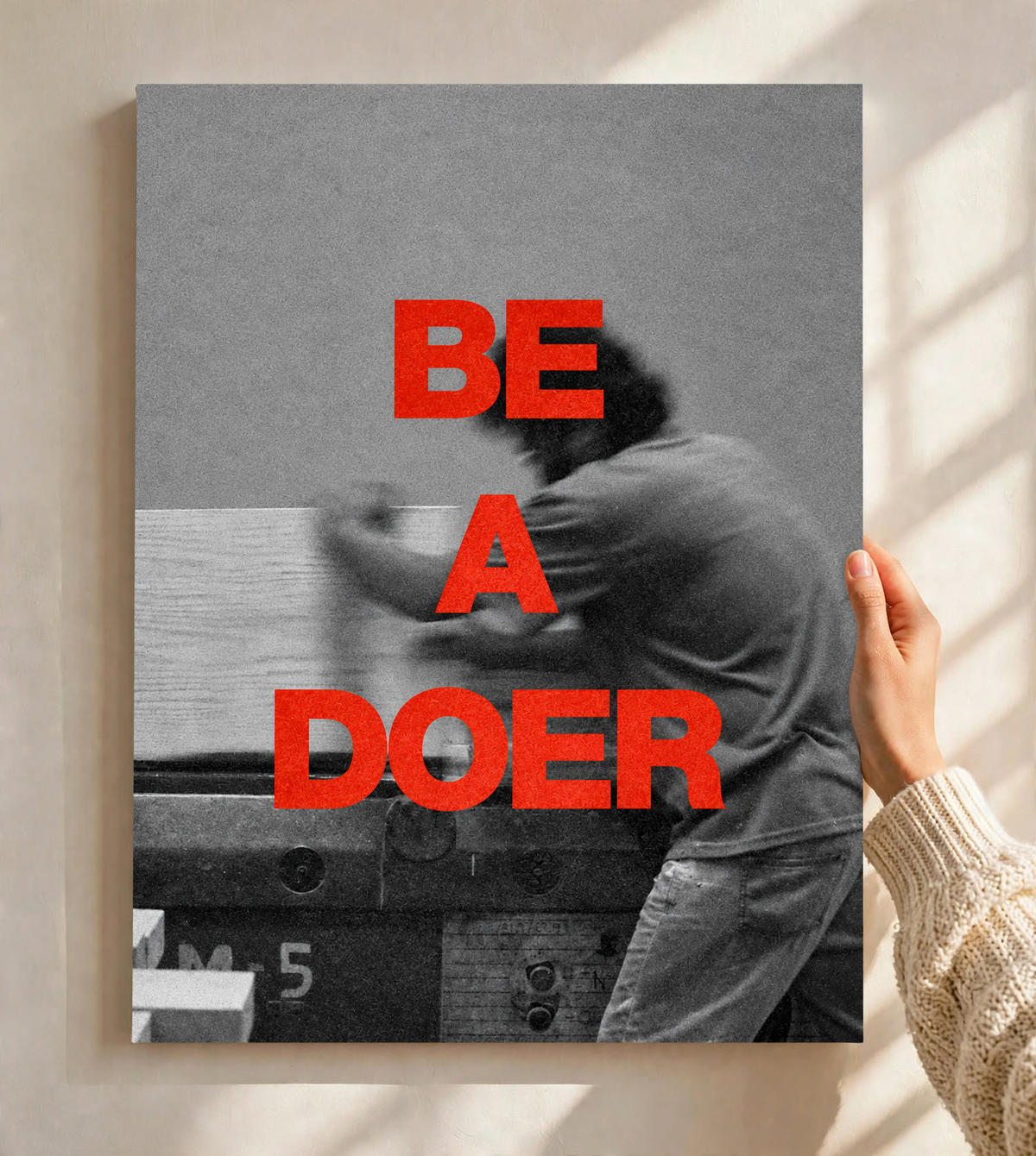 Red Be a Doer lettering over a motion-blurred monochrome figure as an unframed poster held against a pale sunlit wall