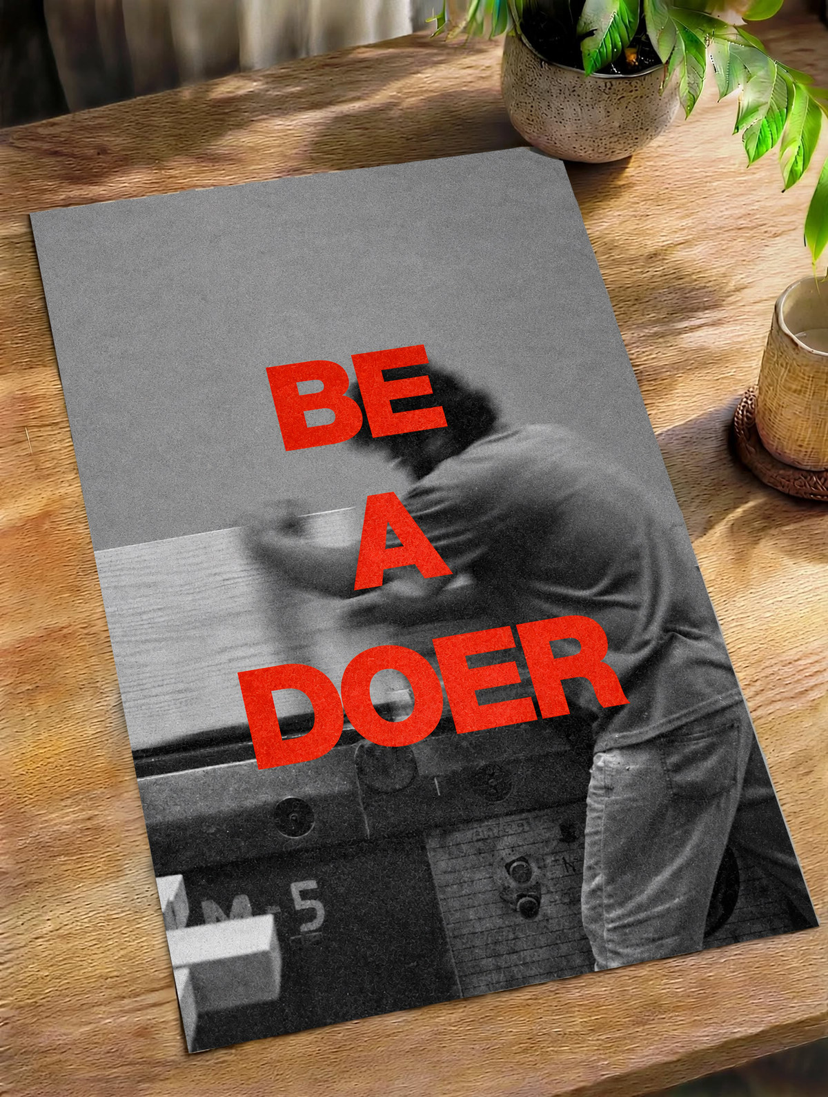 Red Be a Doer lettering over a motion-blurred monochrome figure as an unframed poster lying on a wooden table beside a plant
