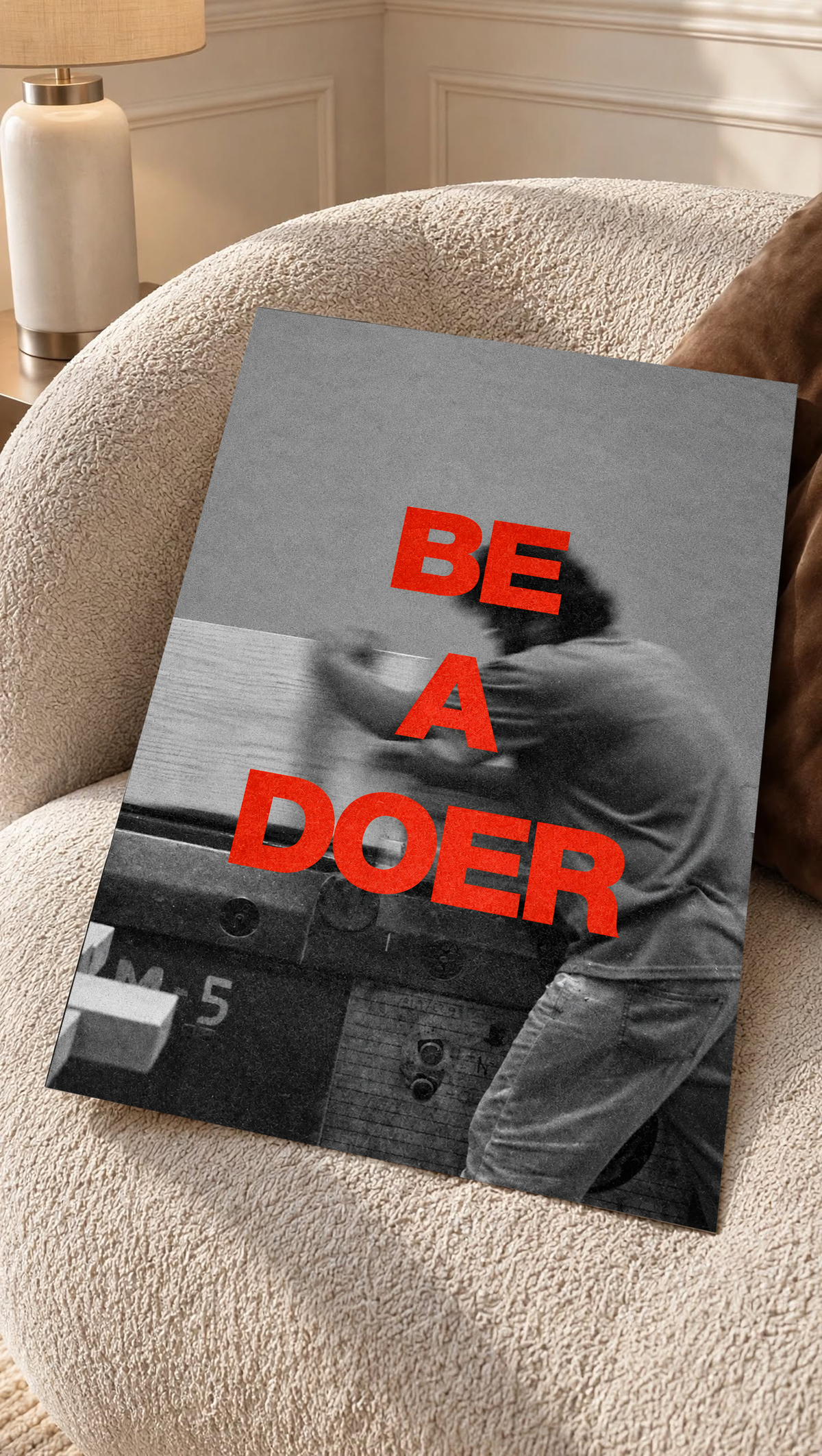 Red Be a Doer lettering over a motion-blurred monochrome figure as an unframed poster resting on a cream woven chair