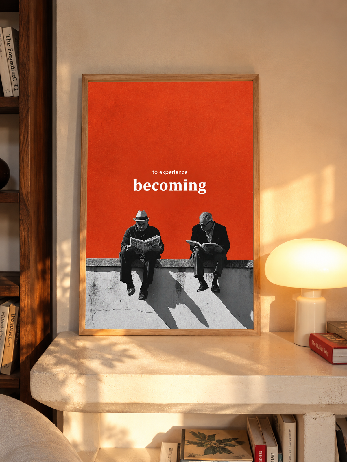 Two monochrome newspaper readers on a ledge beneath Becoming lettering on orange in an oak frame above a pale wooden sideboard beside a glowing lamp