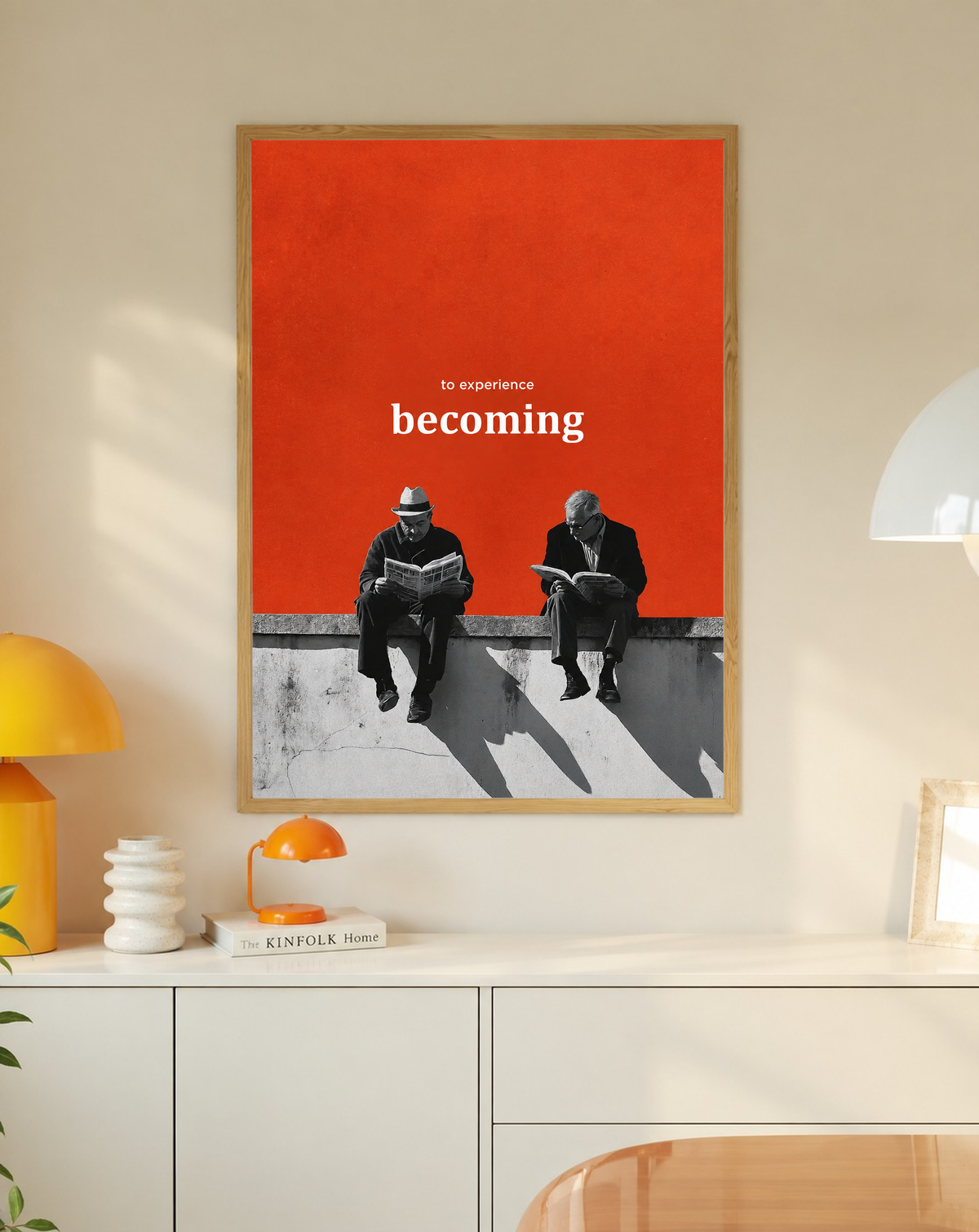 Two monochrome newspaper readers on a ledge beneath Becoming lettering on orange in a slim oak frame on a cream wall above a white cabinet