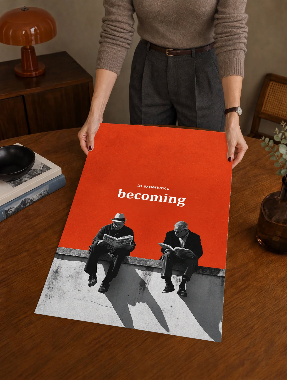 Two monochrome newspaper readers on a ledge beneath Becoming lettering on orange as a large unframed poster held above a wooden desk