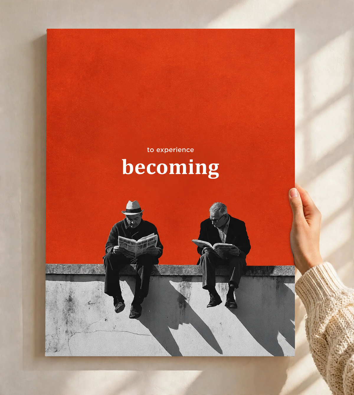 Two monochrome newspaper readers on a ledge beneath Becoming lettering on orange as an unframed poster held against a pale sunlit wall
