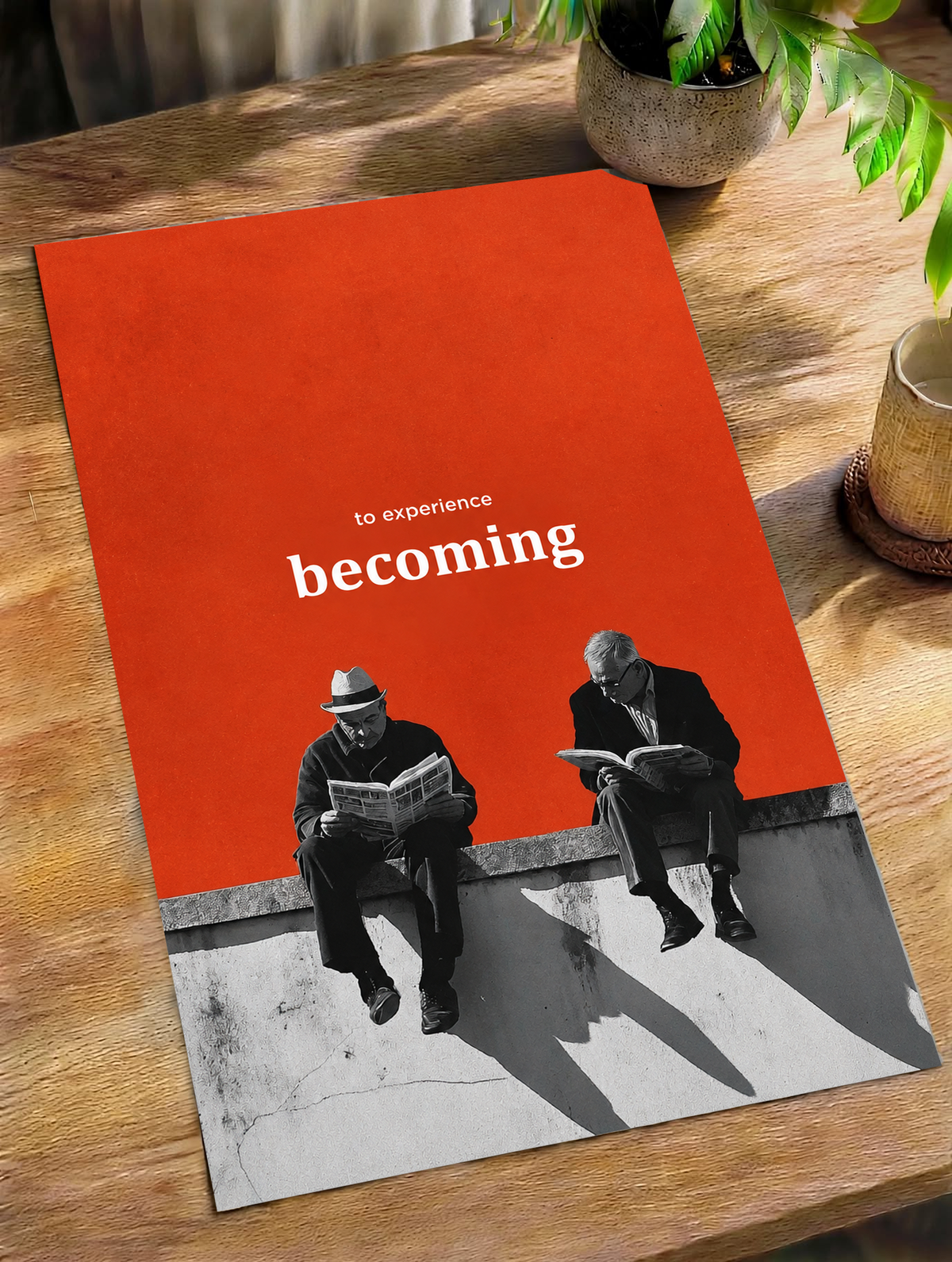 Two monochrome newspaper readers on a ledge beneath Becoming lettering on orange as an unframed poster lying on a wooden table beside a plant