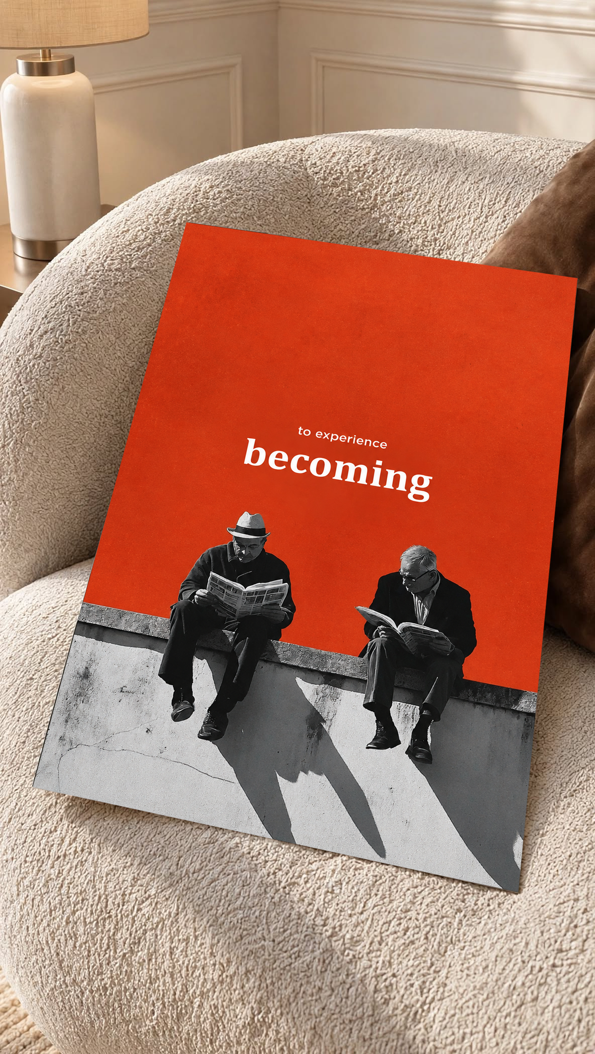 Two monochrome newspaper readers on a ledge beneath Becoming lettering on orange as an unframed poster resting on a cream woven chair