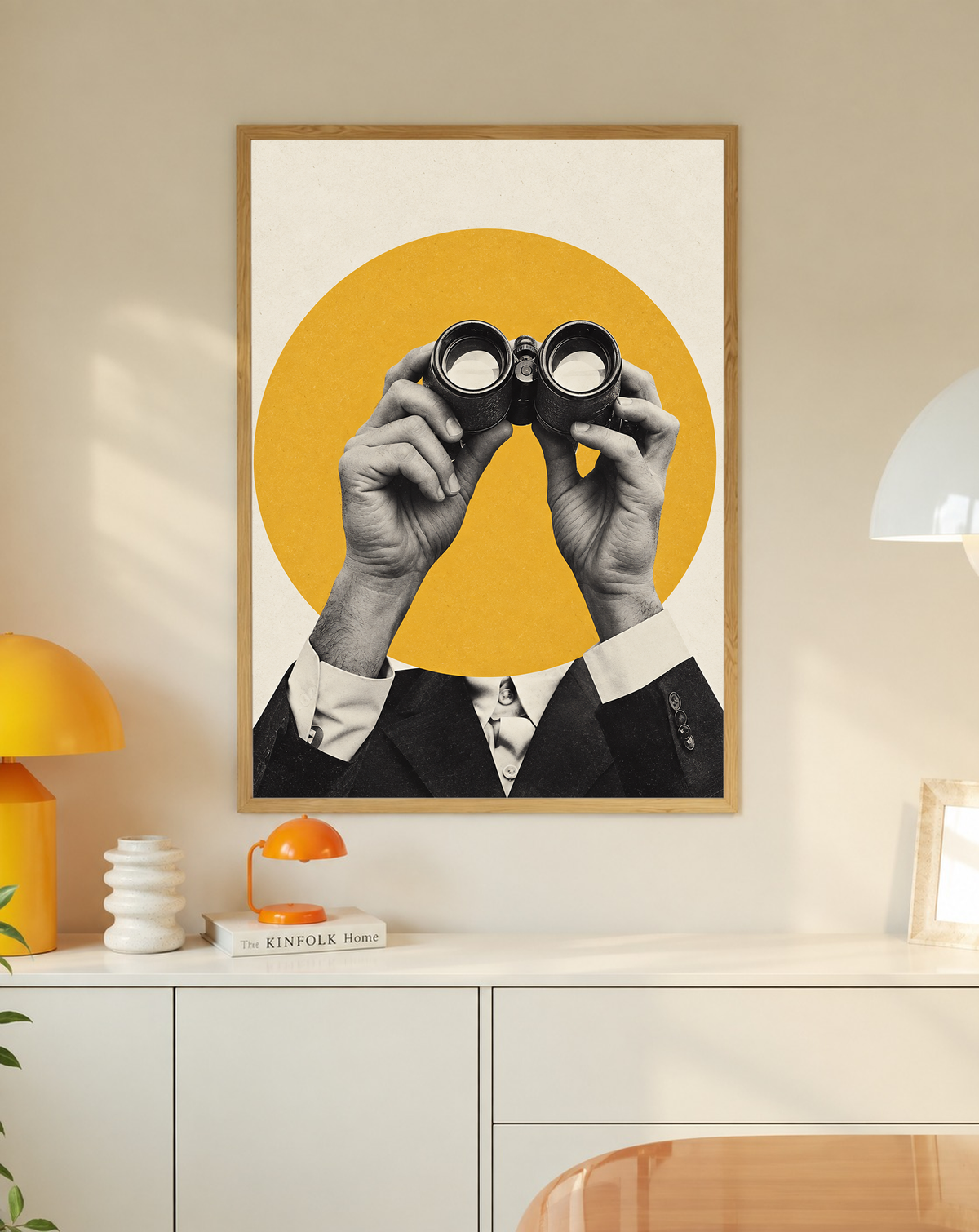 Monochrome hands holding binoculars before a mustard-yellow circle in a slim oak frame on a cream wall above a white cabinet