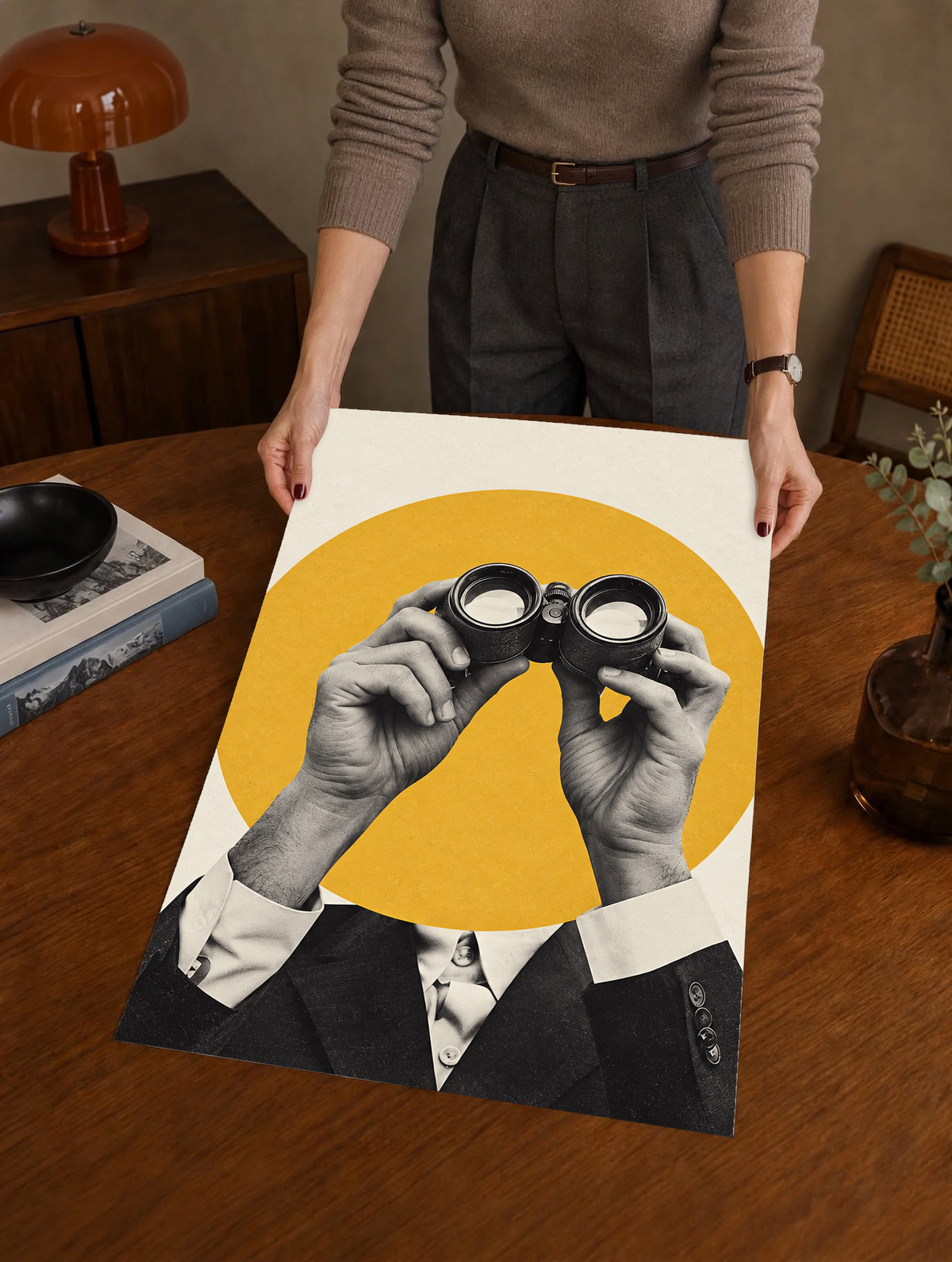 Monochrome hands holding binoculars before a mustard-yellow circle as a large unframed poster held above a wooden desk