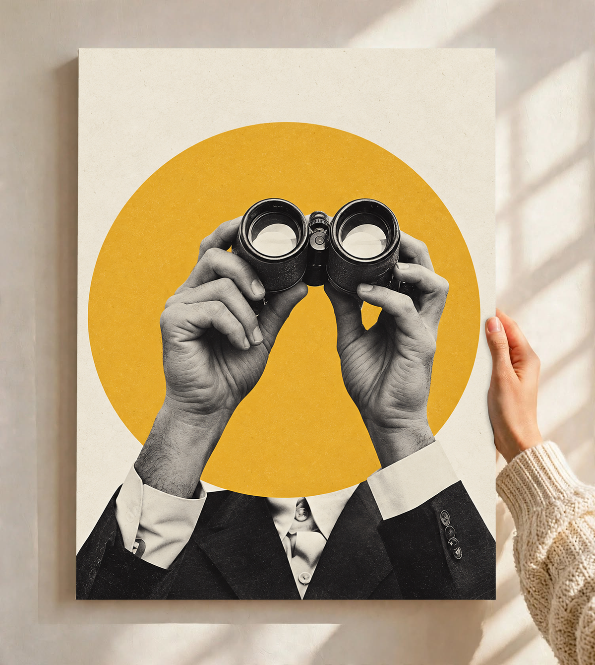 Monochrome hands holding binoculars before a mustard-yellow circle as an unframed poster held against a pale sunlit wall