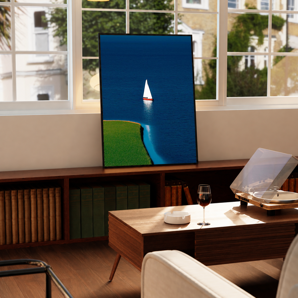 Blue Sailboat Shoreline Print in a black frame near a window and wooden table in a sunlit sitting room.