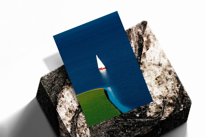 Unframed artwork depicting solitary sailboat on deep blue water beside a green shoreline, presented diagonally on rough stone.