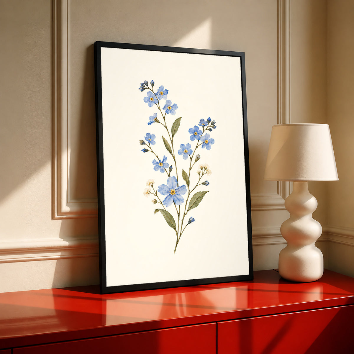 Blue wildflower sprig with small white flowers and olive green leaves in a black frame on a red cabinet beside a cream lamp