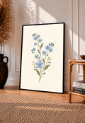 Blue wildflower sprig with small white flowers and olive green leaves in a black frame leaning on a sunlit wooden floor beside books and a rattan seat