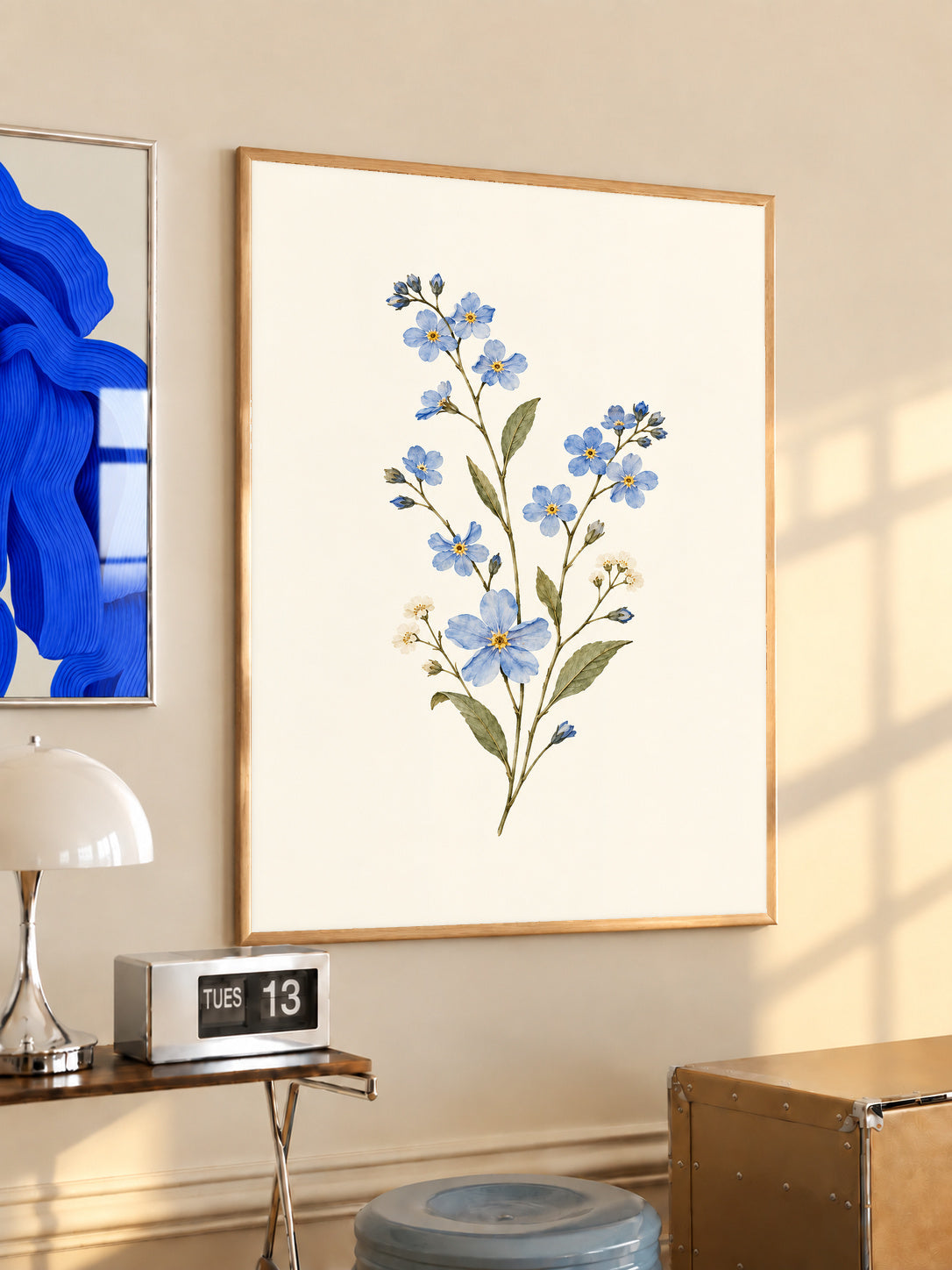Blue wildflower sprig with small white flowers and olive green leaves in an oak frame above a console beside a blue print and calendar
