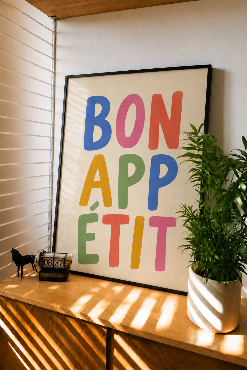 Black-framed BON APP ETIT wall print on a wooden ledge, colorful stacked lettering