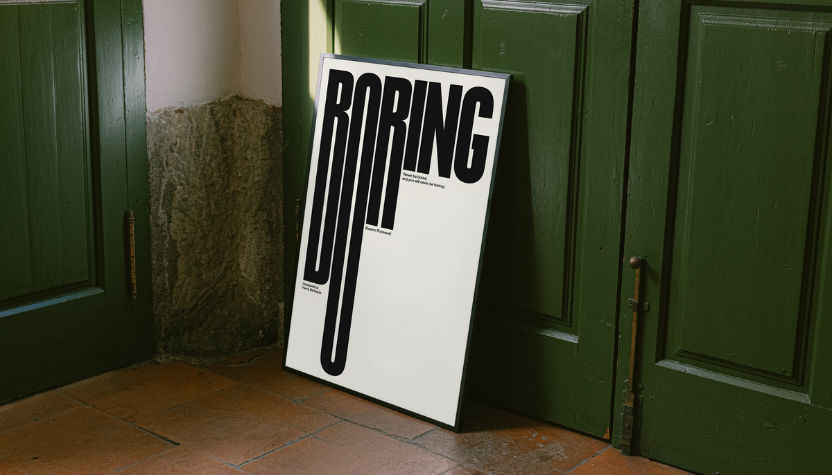 Boring typography artwork with tall black condensed letters and a small quote on cream, styled in a black frame against dark green paneled doors above orange floor tiles