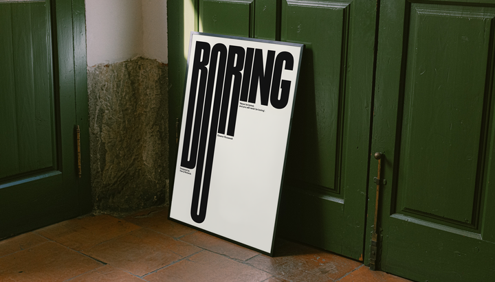 Boring typography artwork with tall black condensed letters and a small quote on cream, styled in a black frame against dark green paneled doors above orange floor tiles