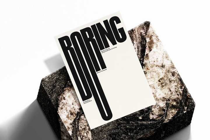 Boring typography artwork with tall black condensed letters and a small quote on cream, shown as an unframed print tilted on a dark stone display against a light background