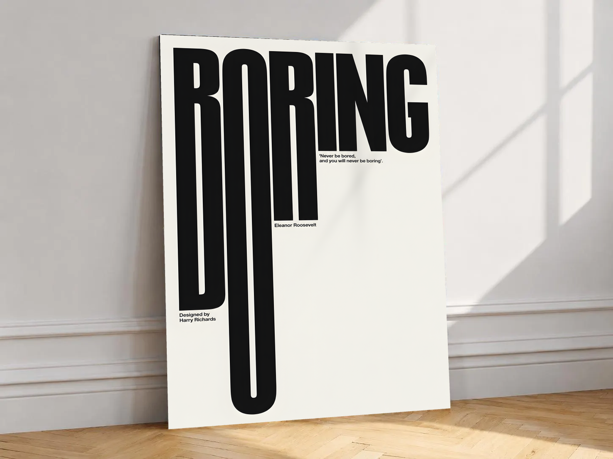 Boring typography artwork with tall black condensed letters and a small quote on cream, shown unframed on a white wall above parquet flooring with a diagonal sun shadow