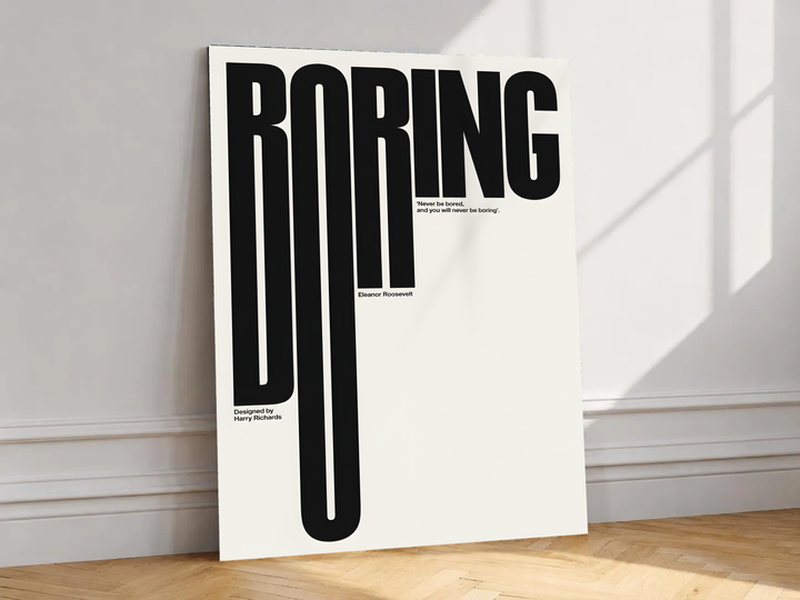 Boring typography artwork with tall black condensed letters and a small quote on cream, shown unframed on a white wall above parquet flooring with a diagonal sun shadow